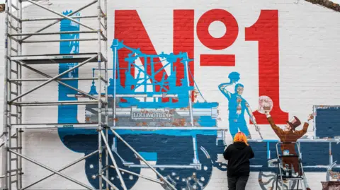 Jess McGowan can be seen painting the bottom of a large mural of a steam train. The words 'No 1' can be seen painted in red above a beautifully captured blue train. The whole thing is painted on a brick wall that has been painted white.