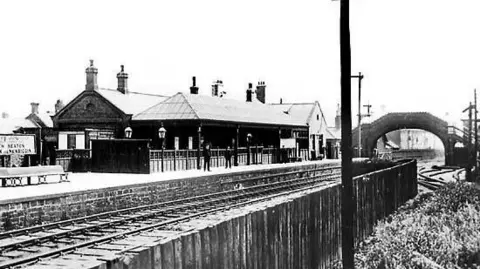 John Mann Collection A black and white photograph of Bedlington railway station with a bridge over the line. Two buildings are visible and there are two people standing on the platform.