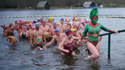 Lots of people with colourful swim caps on a lake being led by a woman dressed with a Christmas tree swimming costume and head display