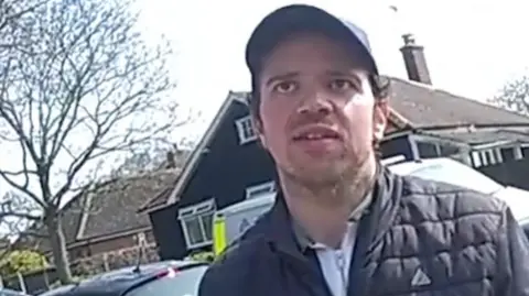 Bodycam image of Luke D'Wit being arrested. He's wearing a hat and padded jacket