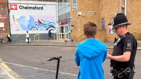 A man in a bright blue tracksuit with his back to the camera. He stands next to an electric scooter and a police man. in the background is a building with the british rail logo and the name "Chelmsford" next to it.