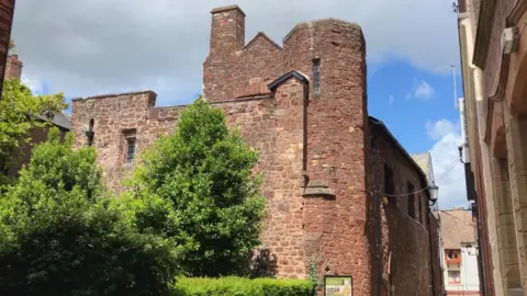 An ancient red brick monastery type building, with trees and bushes in the foreground 