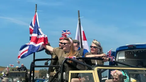 BBC Men and women riding in a car during the Liberation Day 2024 cavalcade. They are wearing sunglasses, khaki outfits and are waving Union Jack flags.