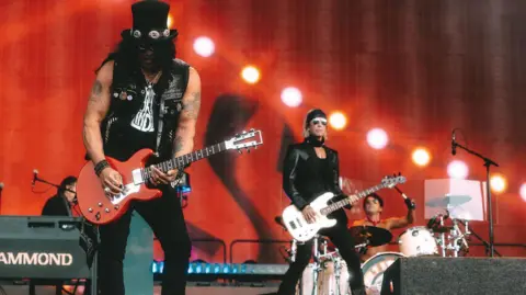 Guns N' Roses A man with a top hat, sunglasses and a black T-shirt on stage playing a guitar. Behind him is another man, also wearing black and sunglasses, playing a bass guitar. In the background is a drummer and a keyboard player. The stage backdrop is orange.