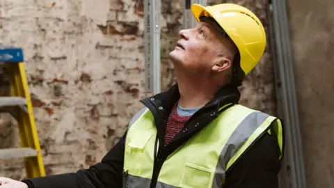 Margarita Hope/True North Matt Moore is a man in his sixties wearing a hard hat and a hi-vis jacket. He is standing inside a building which is being renovated but only bare walls are visible. He is looking up and there is a yellow ladder on the wall next to him 