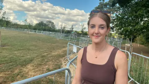 A picture of Jasmine Gray wearing a brown tank top. She is standing in front of a fenced-off area of grass. She is smiling and she has a pair of shades resting on her head.