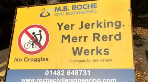 MB Roche A yellow road sign featuring a joke traffic symbol showing a bicycle rider giving a passenger a "croggy" and black letters reading "Yer Jerking, Merr Rerd Werks" (you're joking, more road works)