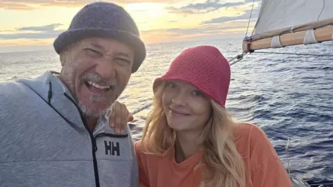 Man with moustache and trimmed beard is wearing a fleece and knitted wool hat. Sitting next to him is a girl with her hair flying in the wind. She has a red wool hat on. She is smiling and they are both on a yacht. 