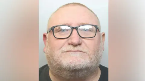 A mugshot of a man with very short blonde hair, a short white beard and black-rimmed glasses. He is wearing a black top.