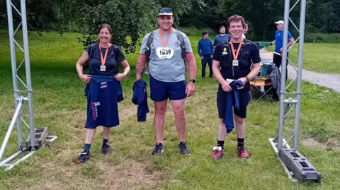 Jamie Price Three runners with square medals on orange ribbons grin under a metal finishing line. On the left is a woman with brown hair tied back in black shorts, t-shirt and trainers. She holds a navy blue t-shirt with 50k in white writing on it. To her right are a taller man in grey t-shit, navy shorts, back trainers, a cap and black sunglasses, and a shorter man with brown hair in back shorts and t-shirt and red trainers. Three other men can be seen behind.