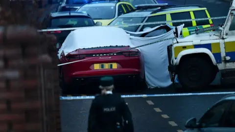 Pacemaker A red Porsche car is covered in a white forensic sheet and surrounded by a white, blue and yellow PSNI Landrover, a green and yellow ambulance vehicle and another emergency response vehicle. A police woman is in the foreground of the image.