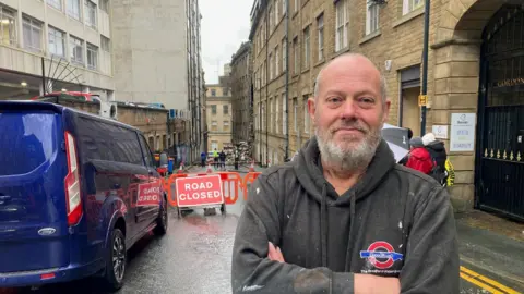 A bald man wearing a dusty black hooded top and folding his arms standing in the street where the building collapsed