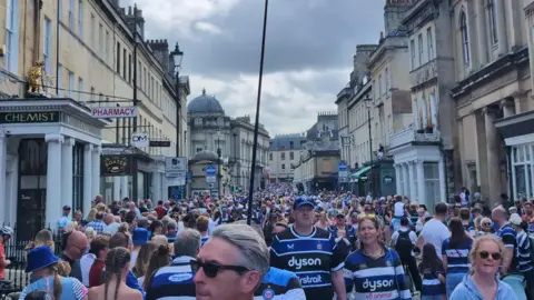 A full city centre street in Bath. Many are wearing Bath rugby shirts.