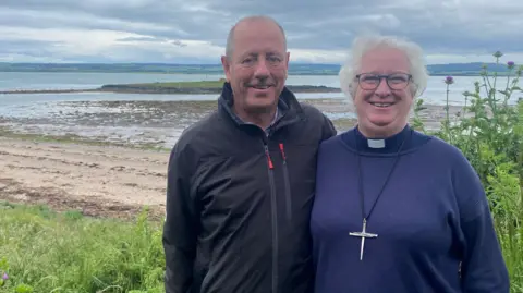 BBC Dr Richard Hills and his wife Dr Sarah Hills are a couple in their sixties. She is wearing a purple top with a clerical collar and a large cross necklace. She has curly grey hair and glasses. He is wearing a zipped-up dark outdoor jacket and has short receding grey hair. Both are smiling. Behind them is a beach with a small low island in the sea and, in the distance, a larger stretch of rolling land across the water.