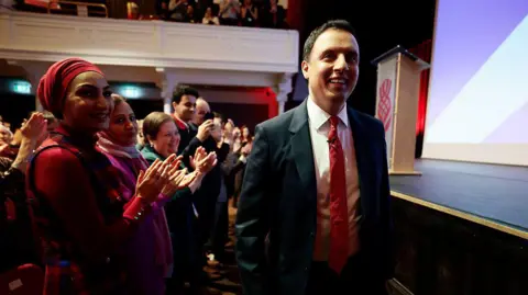 Getty Images Anas Sarwar wears a dark suit with white shirt and red tie. He has short dark hair and walks by the front row of a theatre by the stage. People stand up clapping him 