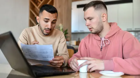 A young couple, two men, look at an electricity bill and a laptop in the kitchen of their home