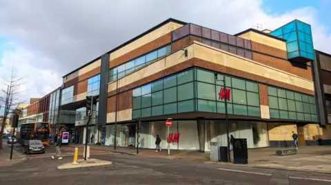 Getty Images An external image of part of the Cabot Circus Shopping Centre in Bristol. The building has flat sides and a flat roof and is a mixture of wood and glass. There is a road running in front of it