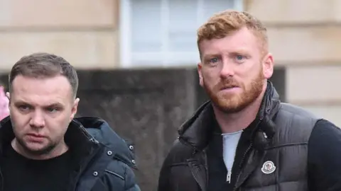 Eddie Lyons, left, who has short, dark hair and is looking slightly off camera. He is wearing a dark jacket over a dark t-shirt. Ross Monaghan, right, is looking straight at the camera. He has ginger hair and a ginger beard and is wearing a dark gilet over another dark top.