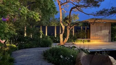 Rebekah Kennington for Asthma + Lung UK A twilight shot of a garden with a tree at the centre of the picture. A path winds past the tree to a wooden structure beyond. More trees and bushes line the path and a wall in the background. A wooden structure stands in front of the back wall on the right hand side of the picture with the path running in front of it. The tree over lookes a pool in the foreground. The trees and the wooden structure are illuminated with soft uplighting.