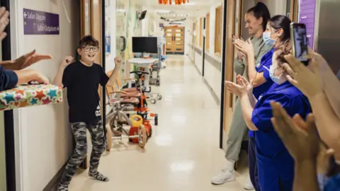 Harley celebrating with his hands in the air while smiling. He is standing in a hospital corridor and is being clapped by staff, who are wearing blue scrubs. Harley has brown hair and is wearing a black T-shirt and camouflage trousers. 