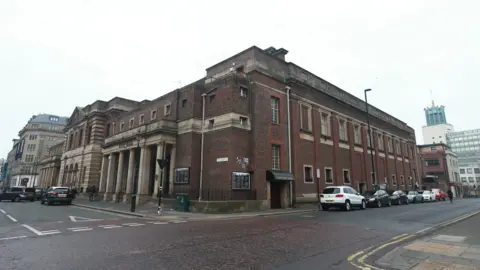 BBC A picture of Newcastle City Hall. The photograph is taken from the corner of the street so two sides of the red-brick building are in view. The entrance stands to the left and has large sandstone pillars in front of the door. The City baths are further to the left.