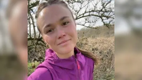 Kit Birks is smiling at the camera, she has brown eyes and  her brown hair pulled back into a ponytail. She is wearing a purple waterproof jacket.