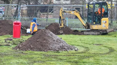 Mark Mather A Digger sedang bekerja di taman bermain dengan pagar besar di belakang 