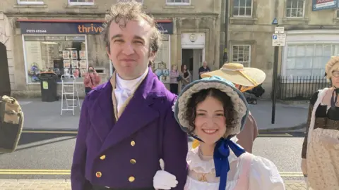 A man and a woman dressed in Regency clothing. He is wearing a purple tunic with gold buttons, and she has a pink and white dress on with a blue frilly bonnet.