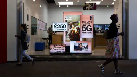 Getty Images Pedestrians walk by a high street branch of Three UK displaying adverts for their 5G network