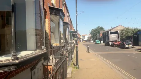 A residential street with a row of shops with black framed windows. There is a pavement next to a two-way street with black lamp posts. There are several cars and a small lorry parked on the road.