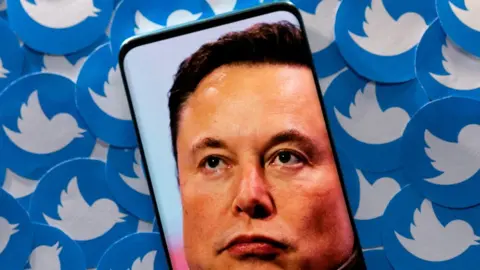 Reuters Elon Musk on phone screen with Twitter logos in the background.