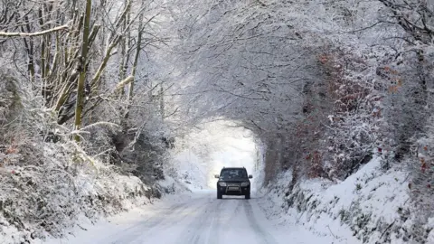 A black car driving in along a snowy road towards the camera. The road is surrounded on either side by trees covered in ice.