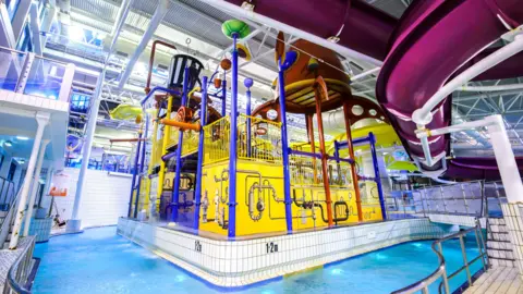 A brightly coloured indoor water park attraction, with yellow, blue and purple colours. A pool of water surrounds it, with flumes and slides visible.