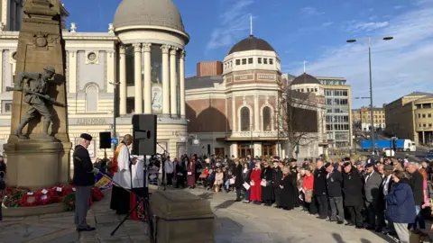 Bradford's Armistice Day service: a crowd of more than 100 people stand in front of the Cenotaph with their heads bowed. A clergyman and a man in military uniform stand directly in front of the monument, next to a large speaker.