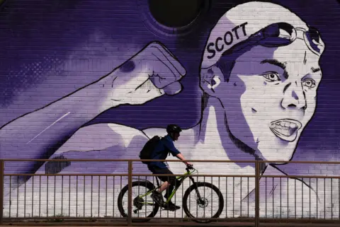 PA Media A man cycles pas a large mural of swimmer Duncan Scott
