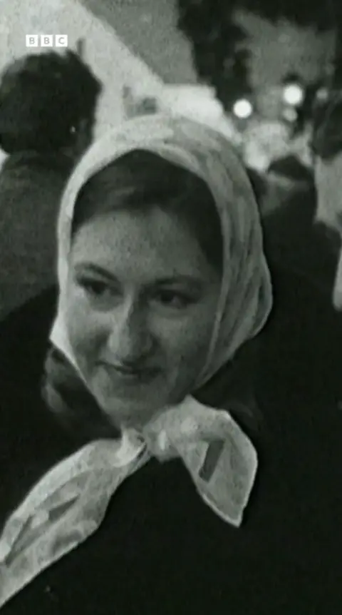 A black and white still from a TV news programme of a woman standing in a shopping street with a few people behind her. It is a head and shoulders shot and she is wearing a silk scarf over her head and smiling.