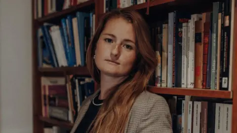 A woman with long brown hair who is wearing earrings and a necklace is standing in front of a book case. 