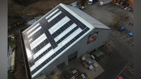 Greg Pilley An aerial view of a modern warehouse building with corrugated grey cladding and a slanted roof, half of which is covered in solar panels. There are large pallets with kegs at the front of the warehouse, and there is a black, white and red Stroud Brewery logo above.