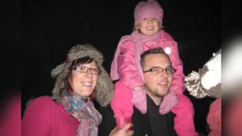 Family handout Jenni Swift - a woman wearing a winter hat with rectangular glasses, a scarf and pink jumper smiling into the camera next to a man with short brown hair also wearing glasses. A young girl wearing all pink including a pink hat is sitting on his shoulders and smiling.