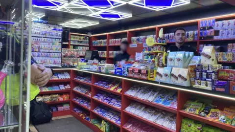 A corner shop counter has several bars of Milka chocolate, Haribo sweets and more on red shelves at the front of the till. A police officer can be seen standing in front of the till, with another one stood behind the till. A man in the middle has been blurred out.