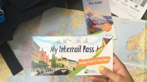 Emily Wyman A traveller holds an Interrail pass next to a map of Europe and a backpack