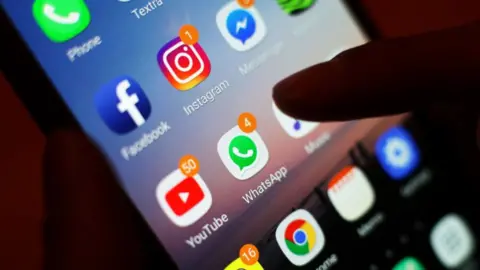 A close-up of a smartphone screen, with a number of social media app icons. A finger is pressing against it.
