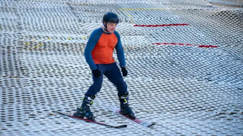 Mendip Activity Centre An older gentleman skiing down an artificial snow slope. He is wearing a helmet, a blue and orange top and black gloves. 
