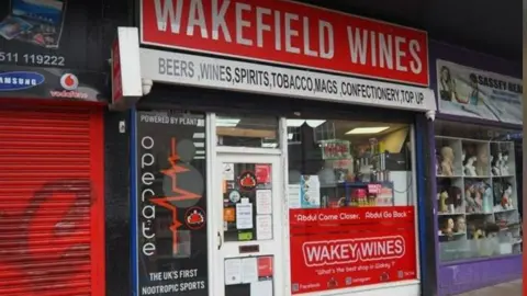 Ernest Wilson Business Agents A red sign has white writing saying 'Wakefield Wines', with a white sign beneath it saying 'beers, wines, spirits, tobacco, mags, confectionery , top up'. Another sign in the window says 'Wakey Wines' and includes the catchphrase 'Abdul come closer, Abdul go back'.