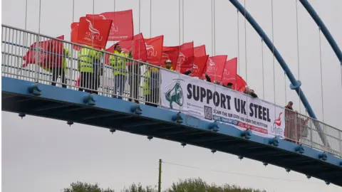 BBC Unite members on bridge waving flags behind sign which reads "support UK steel"