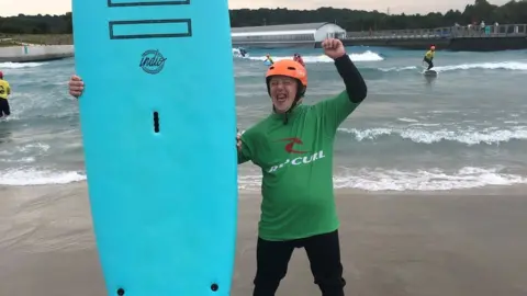 FAMILY HANDOUT Sam wearing a green wet suit, holding up a surf board