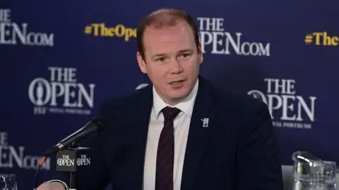 Lyons is sitting in front of a mic talking to the media. The backdrop behind him is for the Open. He's wearing a navy suit with a white shirt and a tie. A jug of water is sitting on the table next to him.