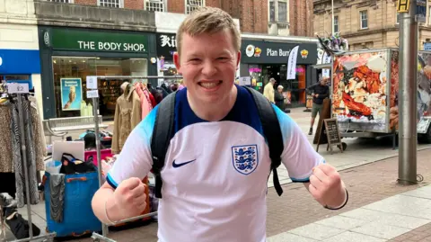 Henry Godfrey-Evans/ BBC Aaron pumping both fists and smiling at the camera. He is wearing a blue and white England football jersey and is standing in the high street, with some market vendors behind him, and the high street chains lining the street.