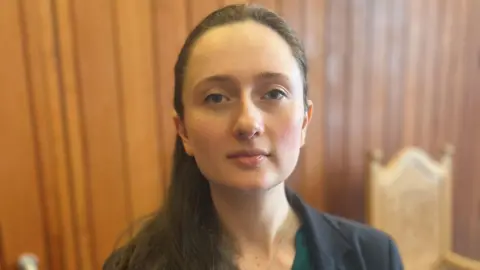 Woman - council leader Heather Woodbridge - looking at camera, with dark hair tied back, wearing a green top and a dark jacket