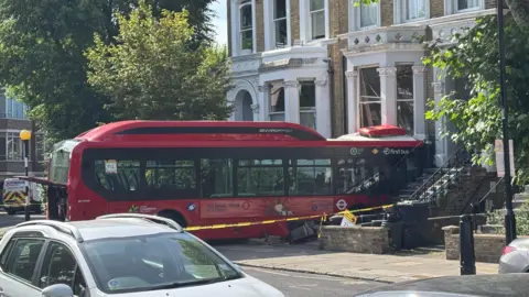 A red bus is seen crashed into a building, with a taped off cordon surrounding it.